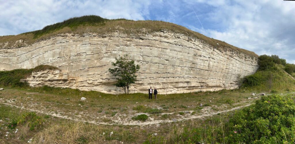 Two people stand in front of a cliff-like bryozoan mound, about 100 feet tall. There's a small tree to the left of the people, and a small foot trail that spans below the cliff.