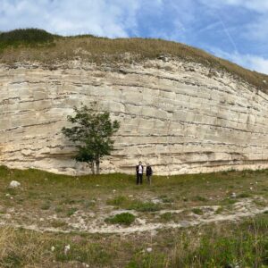 Two people stand in front of a cliff-like bryozoan mound, about 100 feet tall. There's a small tree to the left of the people, and a small foot trail that spans below the cliff.