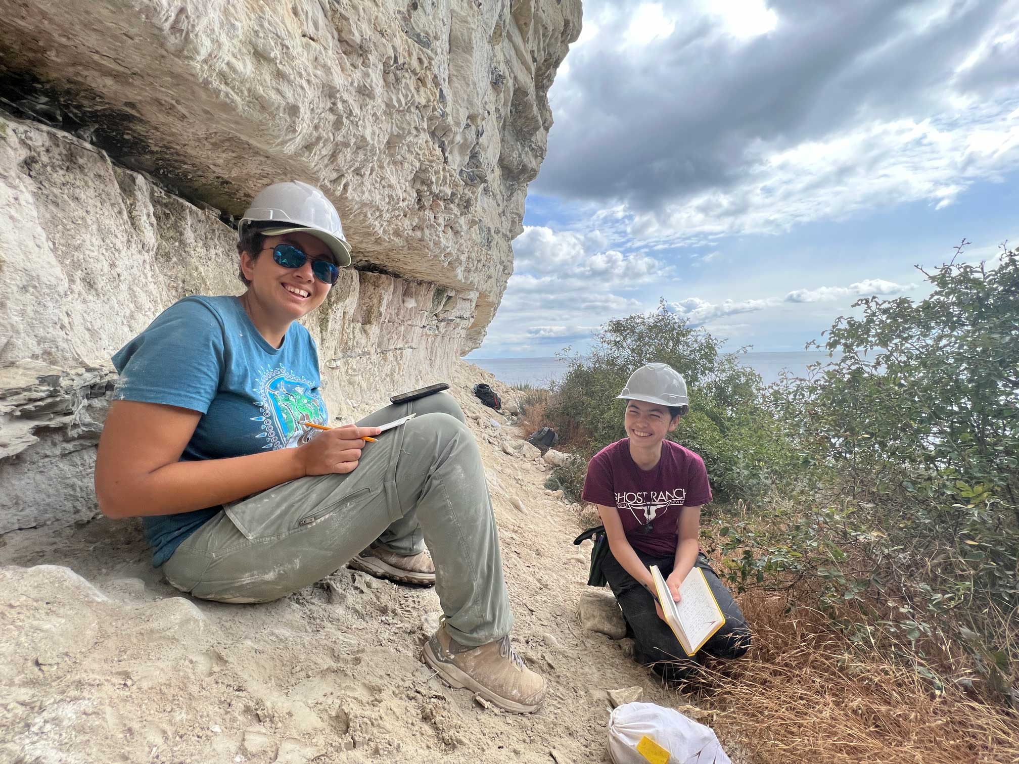Maya is on the left, sitting on some rock formations against a cliff. She is wearing a hard hat, sunglasses, looking at the camera, holding a pencil with a notebook on her lap. Leah is on the right smiling looking at Maya. She is kneeling against the ground holding an open notebook.