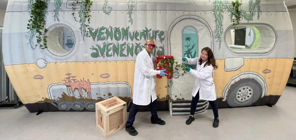 Two people in white lab coats and blue gloves stand in front of an inflatable structure that resembles a stylized van with "VENOMventure aVENENada" written on it. They are holding a potted plant with green foliage between them. The person on the left wears distinctive red glasses while the person on the right has long hair. A wooden shipping crate sits on the floor beside them.